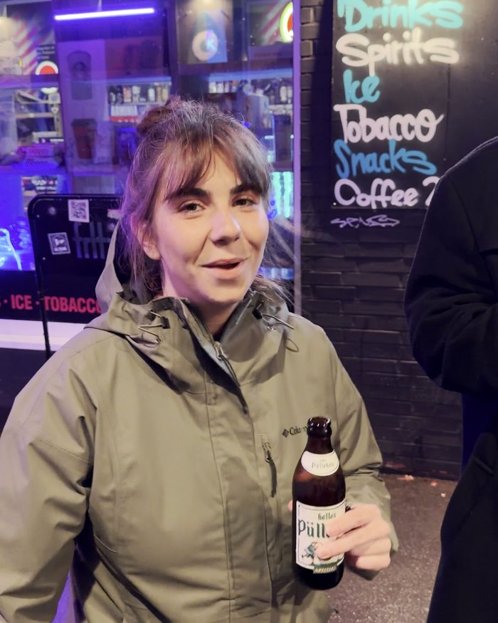 Eine Teilnehmerin mit Bierflasche lächelt vor der Tafel eines Spätis